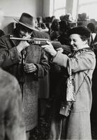 A man wearing a hat and large wool overcoat wipes his nose with a small cloth and smiles. He looks over at the woman next to him who lifts a rifle and closes one eye, pretending to aim.