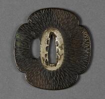 This small, flat metal piece has a quartrefoil shape. Two holes in the middle. Flame-like incision all over the piece. Silver is applied around the center hole.