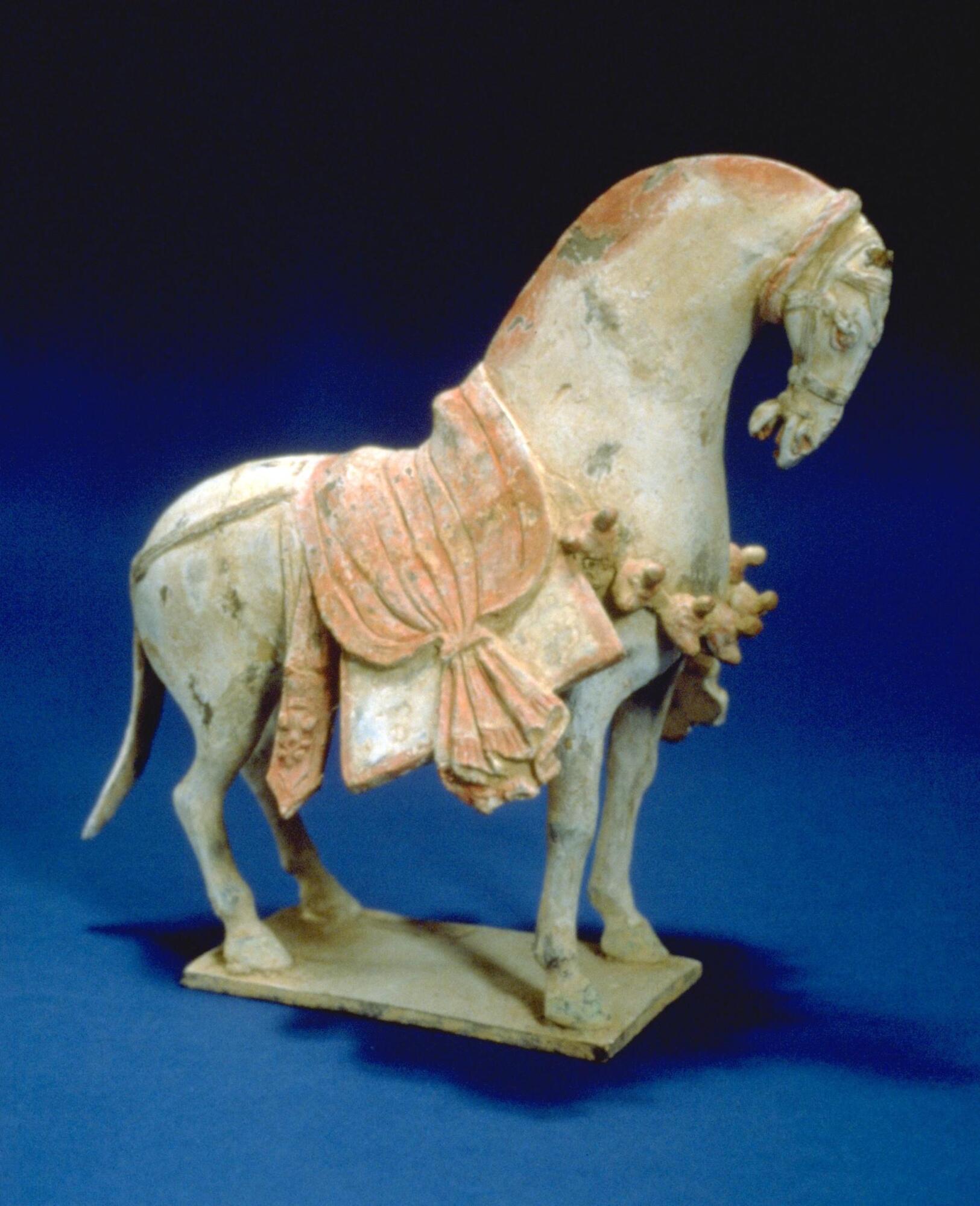 Ceramic figure of a horse standing on a thin ceramic base, which a high-arched neck and a vertical head; large saddle with tassels; traces of orange-ochre, pink red and white pigments