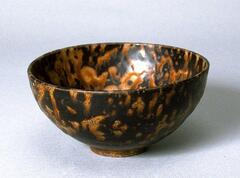 A hemispherical stoneware bowl with a direct rim on a footring, covered in dark brown and amber, tortoise-shell glaze. 