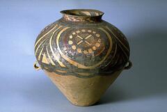 A light reddish-buff earthenware <em>guan </em>罐 jar with a wide globular upper body and conical lower body on a flat base, and a tall narrow neck with everted rim. There are two diametrically opposed lug handles at the waist. The upper half of the body is painted with black and red pigments to depict four whirls of concentric circles, each containing a roundel made of joined circles with an "X" through the center surrounded by a network pattern. The four circular motifs are confined between solid band borders, with a garland border below. Around the neck are bold thick black chevrons.