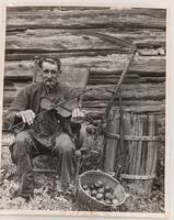 A man smokes a pipe and plays a violin while sitting on a rocking chair outside. Beside him are a barrel, rifle, scythe, and metal bucket of apples.