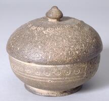 Stoneware jar with natural ash glaze and rounded lid. Along the widest horizontal stretch is an incised circle-and-dot design. A design of concentric circles also loops around just as the rounded curve of the lid begins to flatten into a plateau on which a cintamani style knob rests.