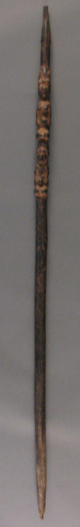 Carved wooden staff topped by a piece of metal with two standing human figures along the top third. The figures appear to be a woman and below her, a man. The bottom of the staff ends in a point. 