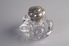 An inkwell made of glass and cur crystal has a special body shape of twisted swirls. There is a round metal lid cover.