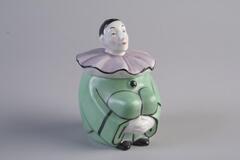 A porcelain inkwell in the shape of a clown. The clown is sitting with his arms around his legs.  He is wearing a green costume with a light purple ruff, trimmed in black.
