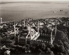 This photograph shows an aerial view of a mosque positioned on a hilltop overlooking a cityscape on a coast.