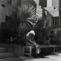 Older man in a suit and cap seated on a bench, his head is down.There are palm trees in the background and the city skyline of buildings.