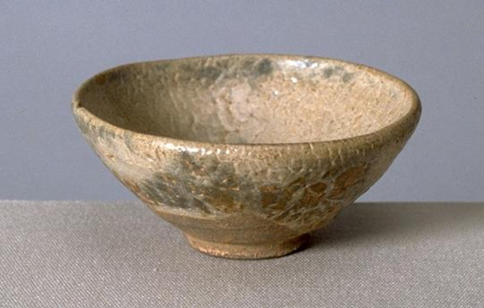 A small round bowl on a tall straight foot ring, made from brown stoneware covered in a thick brown glaze with gray mottling. The too-thick glaze is crawling away from the underlying clay body.