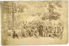 A group of soldiers stand and sit in a landscape; some face the camera while others are posed engaged in conversation. Behind them stand several tall trees and between them a tent with the flaps decorated with an American flag. Seated alone to the left is a young African-American man holding a broom.
