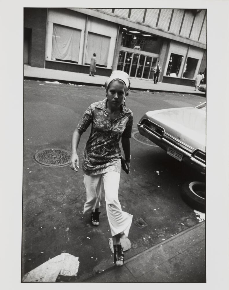 Photograph of a woman wearing a shirt and pants, walking from the street onto the curbside, staring directly at the camera.