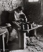 Shows a man leaning over at a potters wheel. The floor and walls appear to be made of dirt.