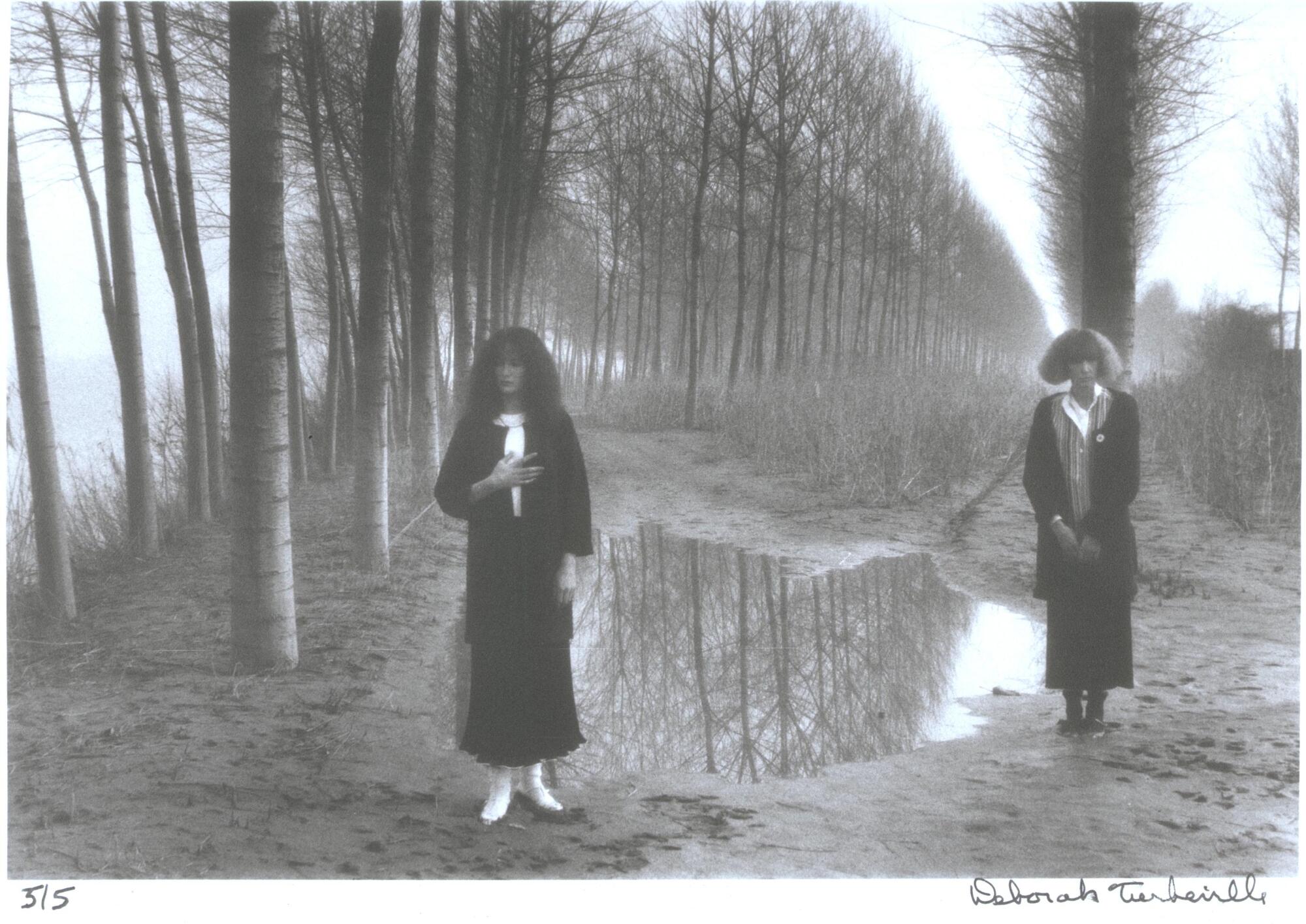 A photograph of two women standing outdoors. They stand next to a puddle of water, one left of center, one on the right side of the frame. Rows of trees extend into the background behind them.