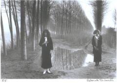 A photograph of two women standing outdoors. They stand next to a puddle of water, one left of center, one on the right side of the frame. Rows of trees extend into the background behind them.