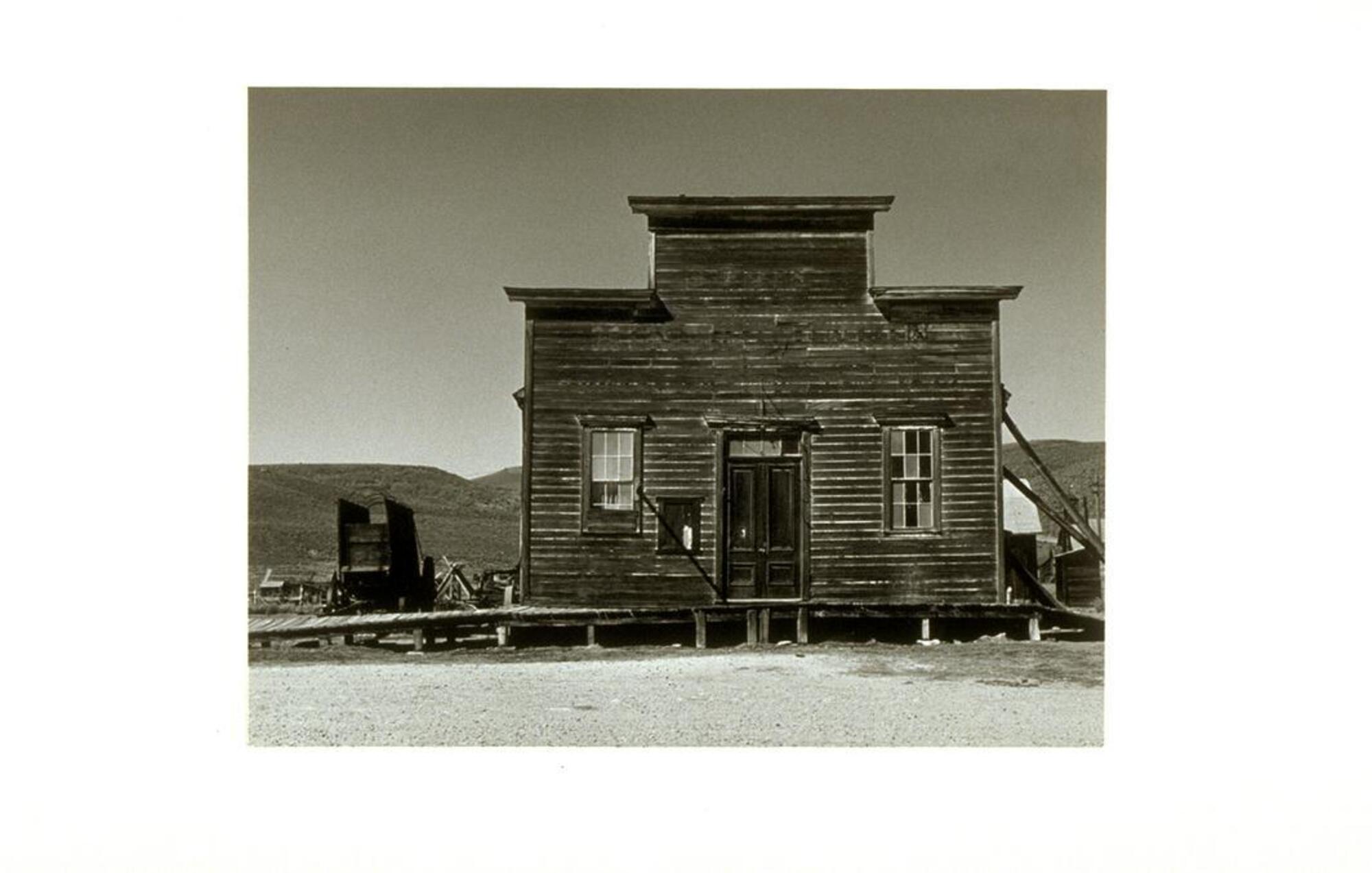 Photograph of a square-shaped wooden building with broken windows and a wagon alongside it.