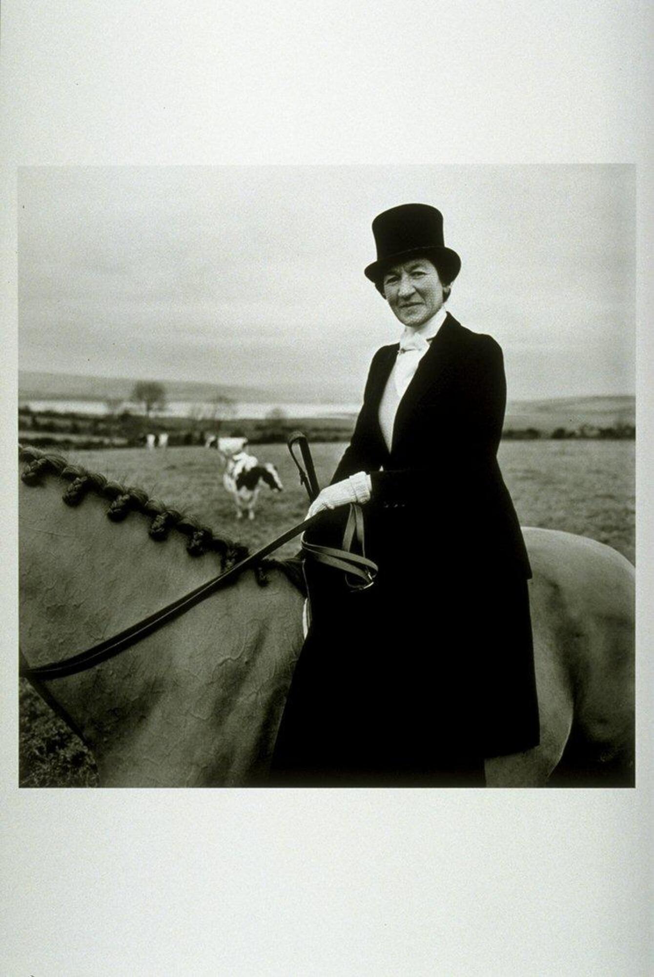 This photograph depicts an elegantly dressed woman on top of a horse in a rural setting.