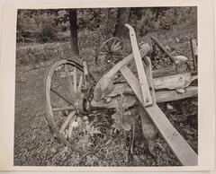The remains of deteriorated farming equipment, included metal-rimmed wheels and wooden handles, sit in front of a wooded area. Plants and flowers have grown among the remains of the equipment.&nbsp;