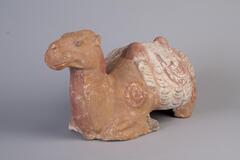 An earthenware bactrian camel seated on bent legs, head raised, with saddle and gear. It is painted with polychrome mineral pigments. 