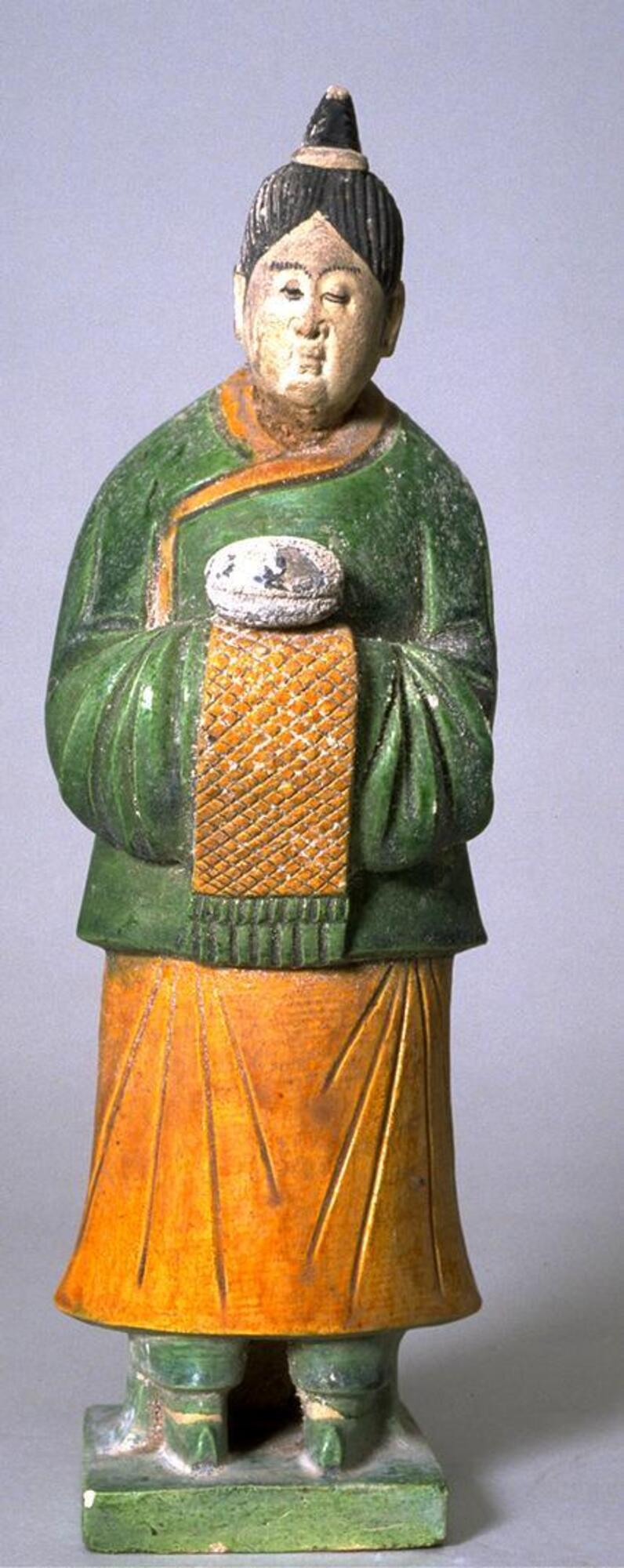 An earthenware female figure with three-color polychrome glazes, wearing a green tunic over a long amber skirt, green trousers and boots on a platform, carrying a round box, her hair pulled up in a top-knot and her face painted with polychrome mineral pigments. 