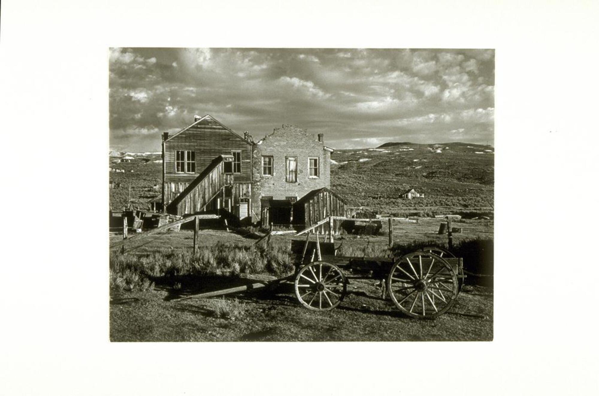 A photograph of two wooden buildings in a western landscape with a wagon in the foreground.