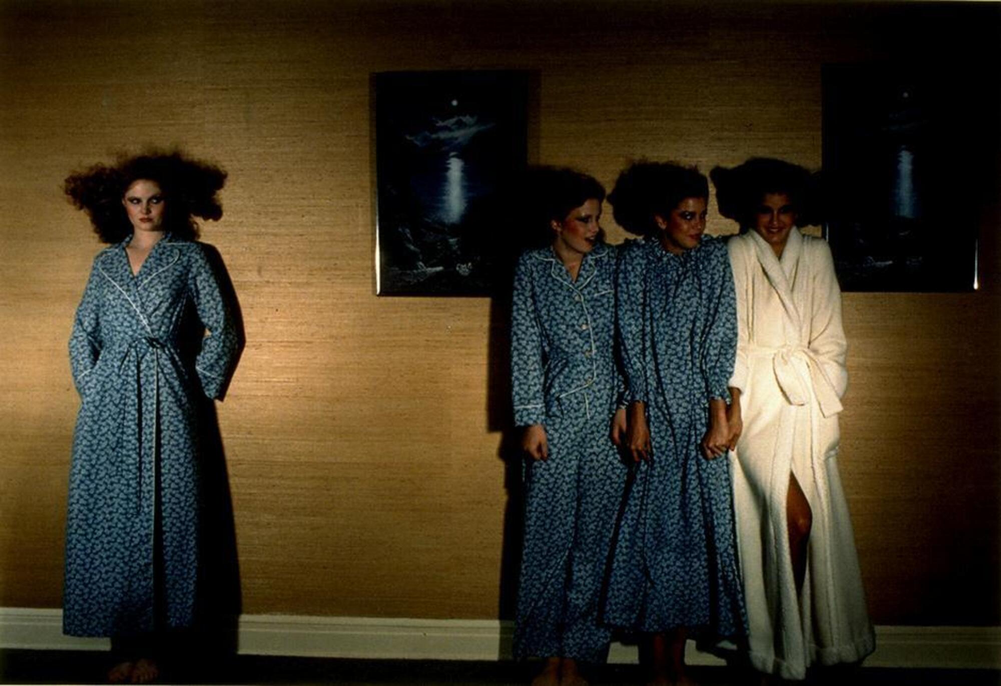 This work is a color photograph of four models standing against a wood-paneled wall. Two identical decorative prints of a night scene hang behind them. On the right, three models are framed by these wall hangings, while another model to the left stands alone and skeptically eyes the happy trio.