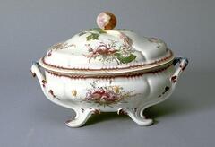This ovoid tureen stands on four curved, leaf-like feet that sprout upward and join together to form a pair of handles at either end of the vessel. The body of the tureen, composed of gentle undulating curves, is decorated with bright red and yellow flowers and leaves painted with overglaze enamel. A scallion, modeled in relief, rests bundled together with sprigs of parsley on top of the lid, introducing a playful trompe-l'oeil element. An onion forms the knob on the top of the lid.