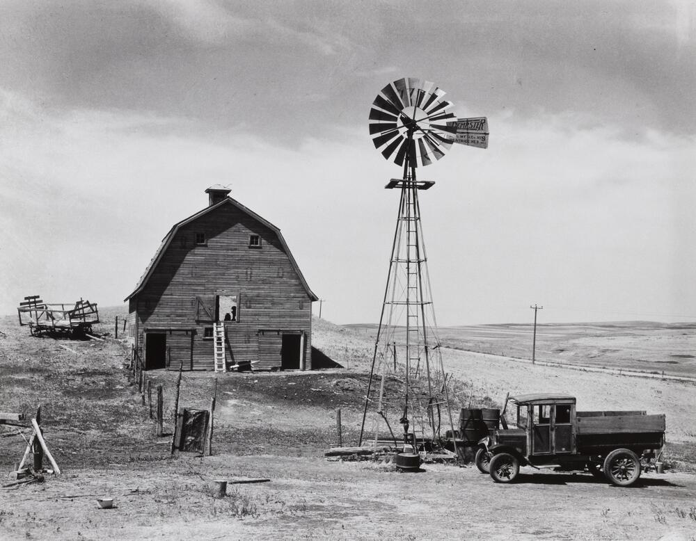A farmstead with a barn, windmill, truck, and hay wagon near a road.