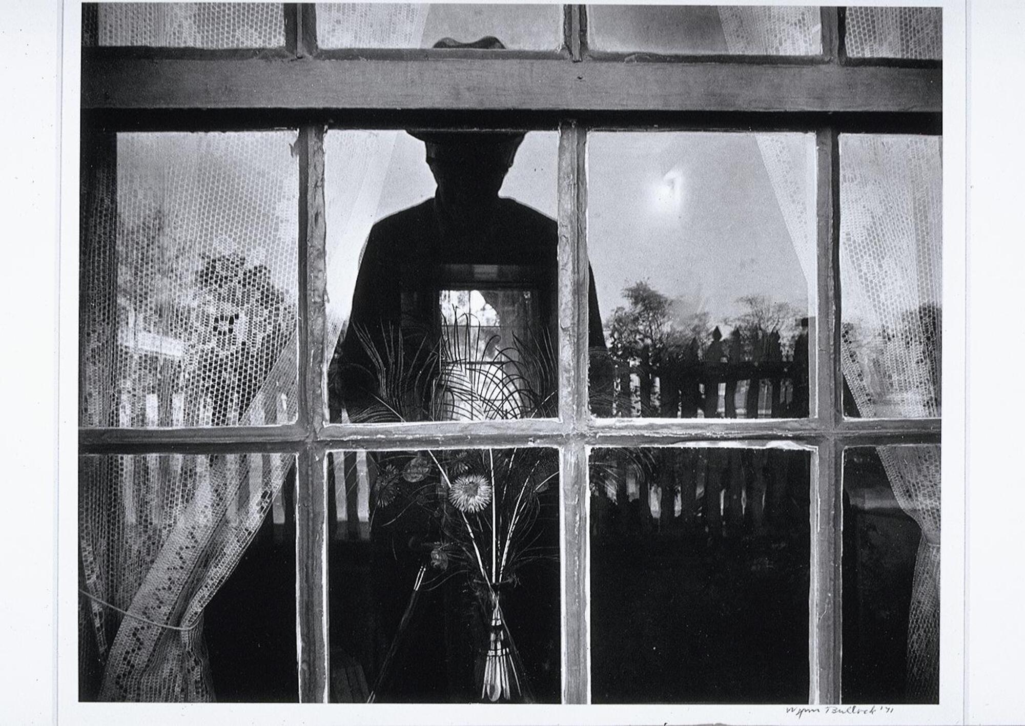 This photograph depicts a window of a house with the photographer’s reflection cast in it. Also visible in the reflection are the white picket fence and trees behind the photographer, as well as objects inside the interior of the house.