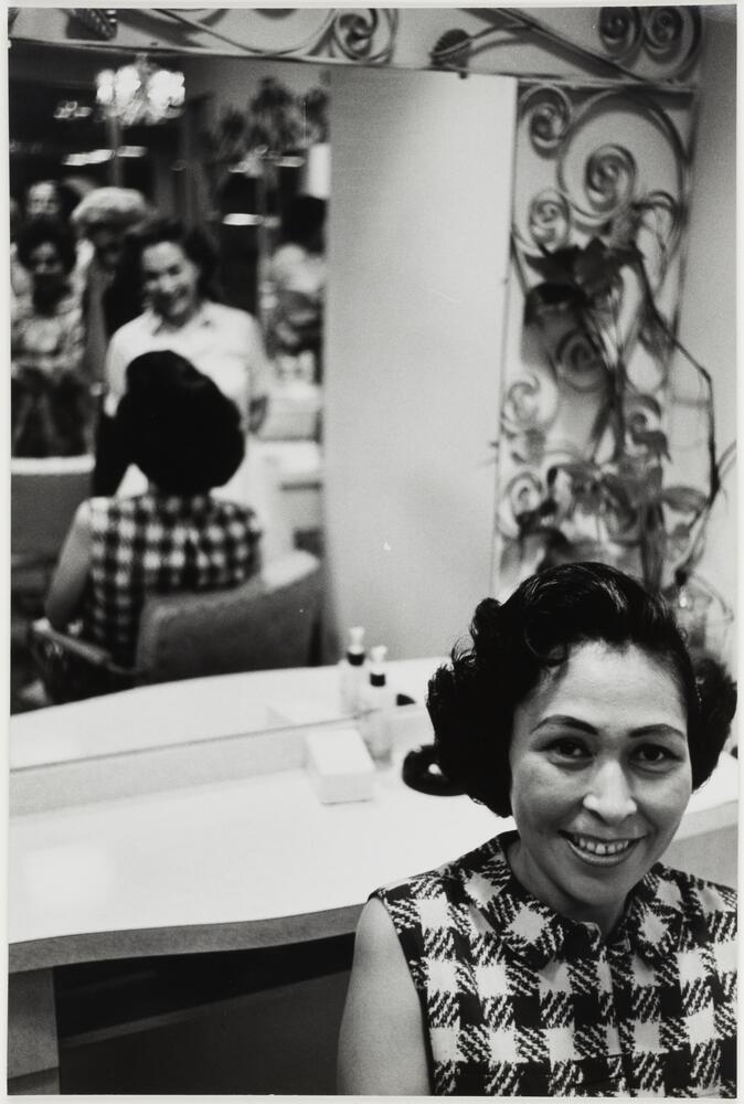 Image of a smiling woman with short, dark hair, seen from the shoulders up in the far right foreground of the frame. A blurred reflection of her and a standing woman in a light dress can be seen in the far left background.