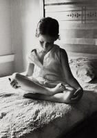 The image shows a young girl sitting cross-legged on a bed, looking down, her hands resting on her knees.