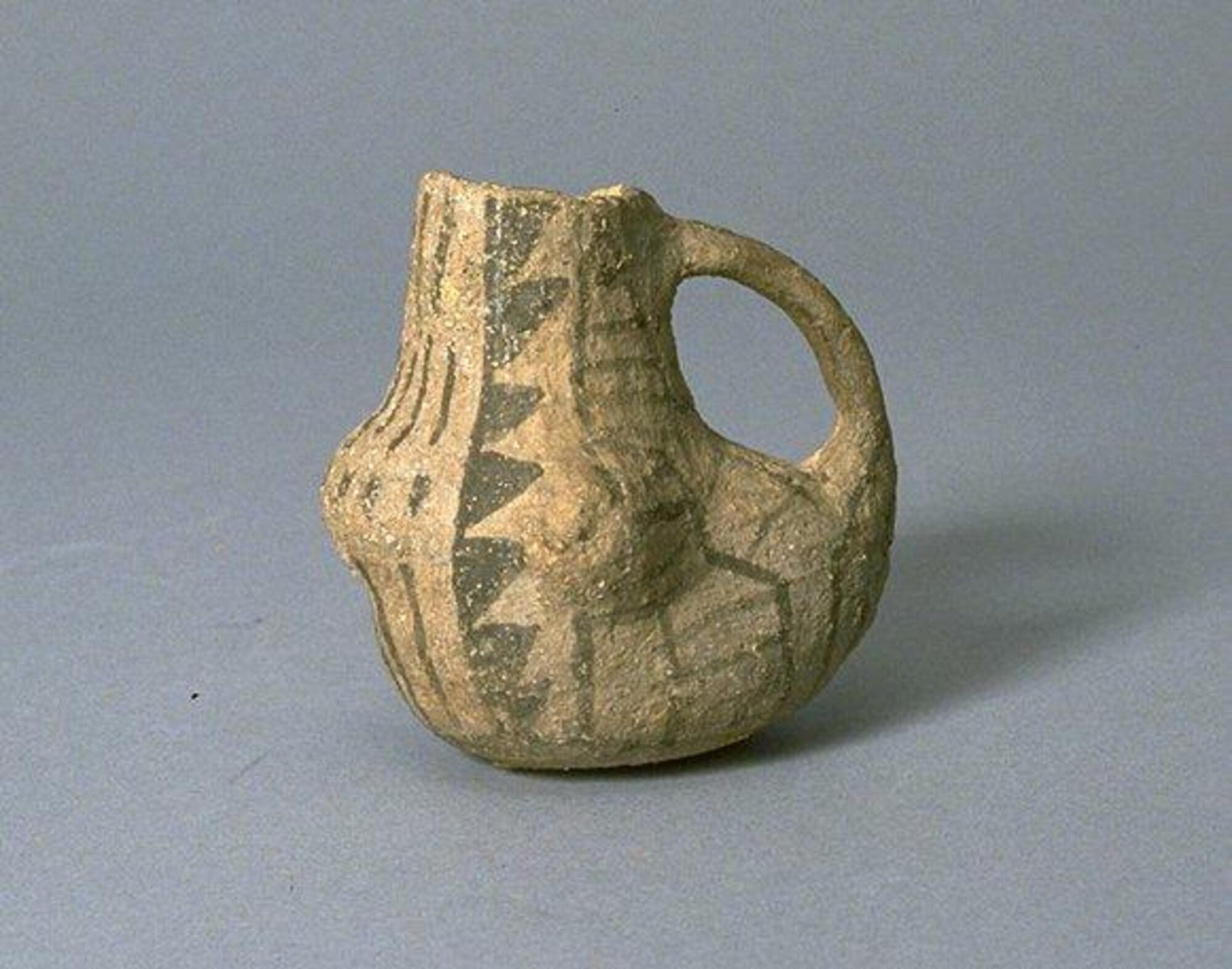 This mug-sized vessel has a bulbous body, a narrower tall neck, and handle. It is beige with black painted geometric and striped design.