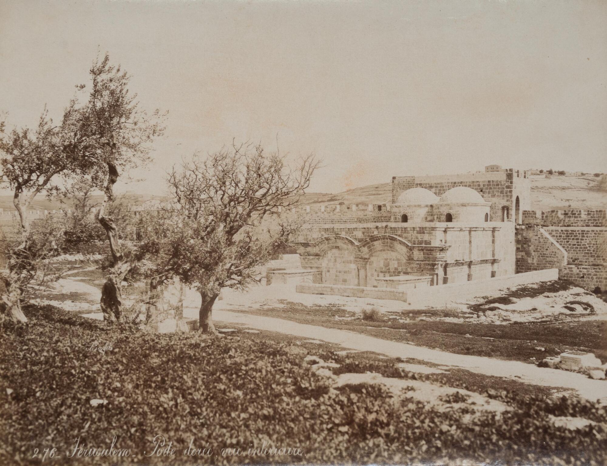 On the right-hand side of the photograph, a rectangular stone structure capped with two domed cylindrical turrets abuts a crenellated stone wall. Three trees in the foreground dominate the left-hand side of the image.