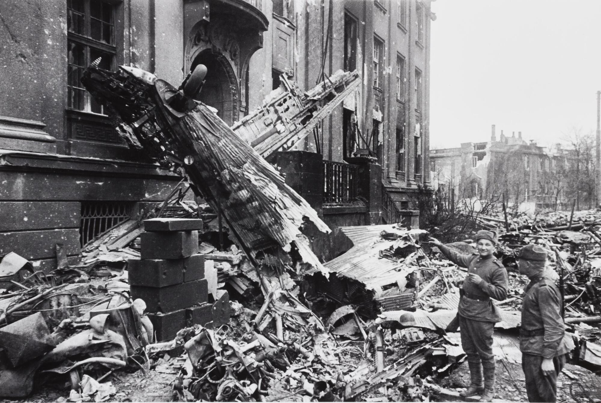 Street scene with two soldiers who look at the remains of a destroyed plane that leans against a bombed-out building fa&ccedil;ade.