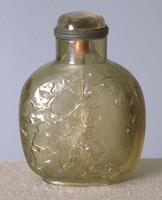 A square shaped quartz snuff bottle with rounded corners. Carved in raised relief are decorations of flowers and leaves. On the top is a stopper.
<div>&nbsp;</div>
