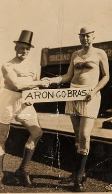 Two men stand in front of a stage, looking at the camera and shaking hands.&nbsp;Both men are wearing a hat, brasserie, and boxer shorts. The men are holding a sign between them that reads &quot;ARON-GO-BRAS.&quot;
