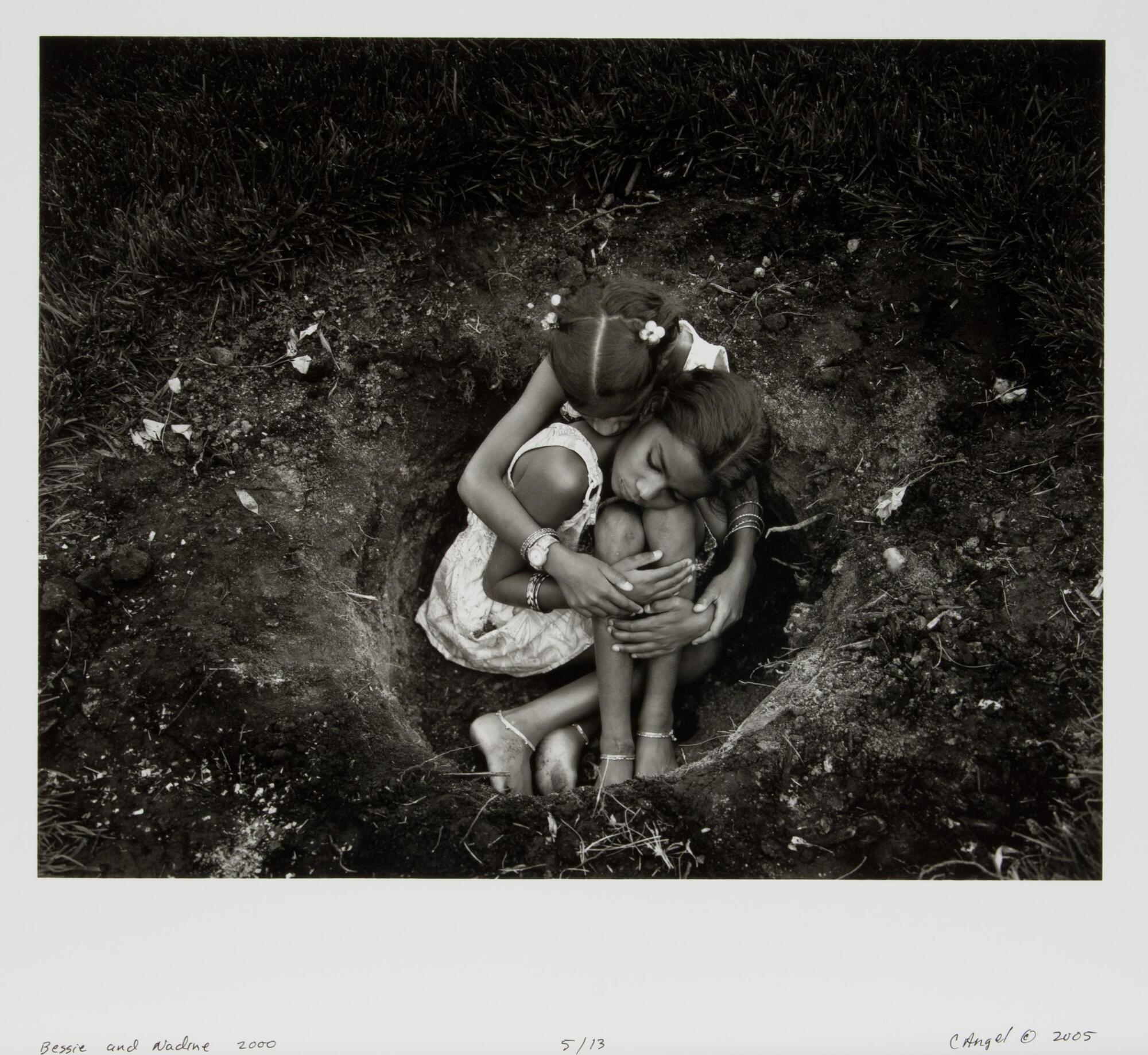 Two girls cuddled in a hole surrounded by dirt.