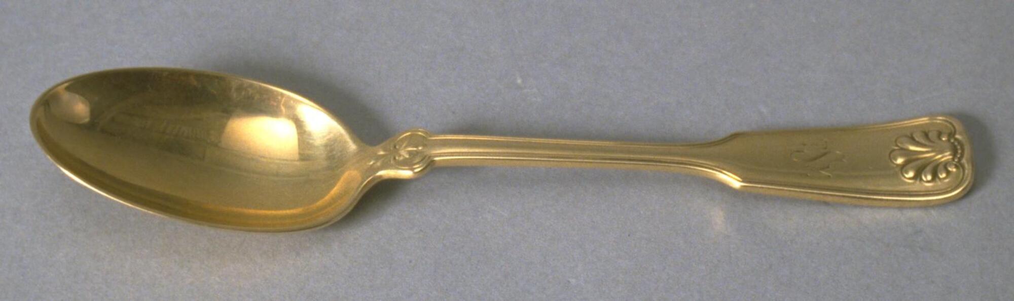 Gold spoon with thin handle that widens at the end with a decorative element at the tip