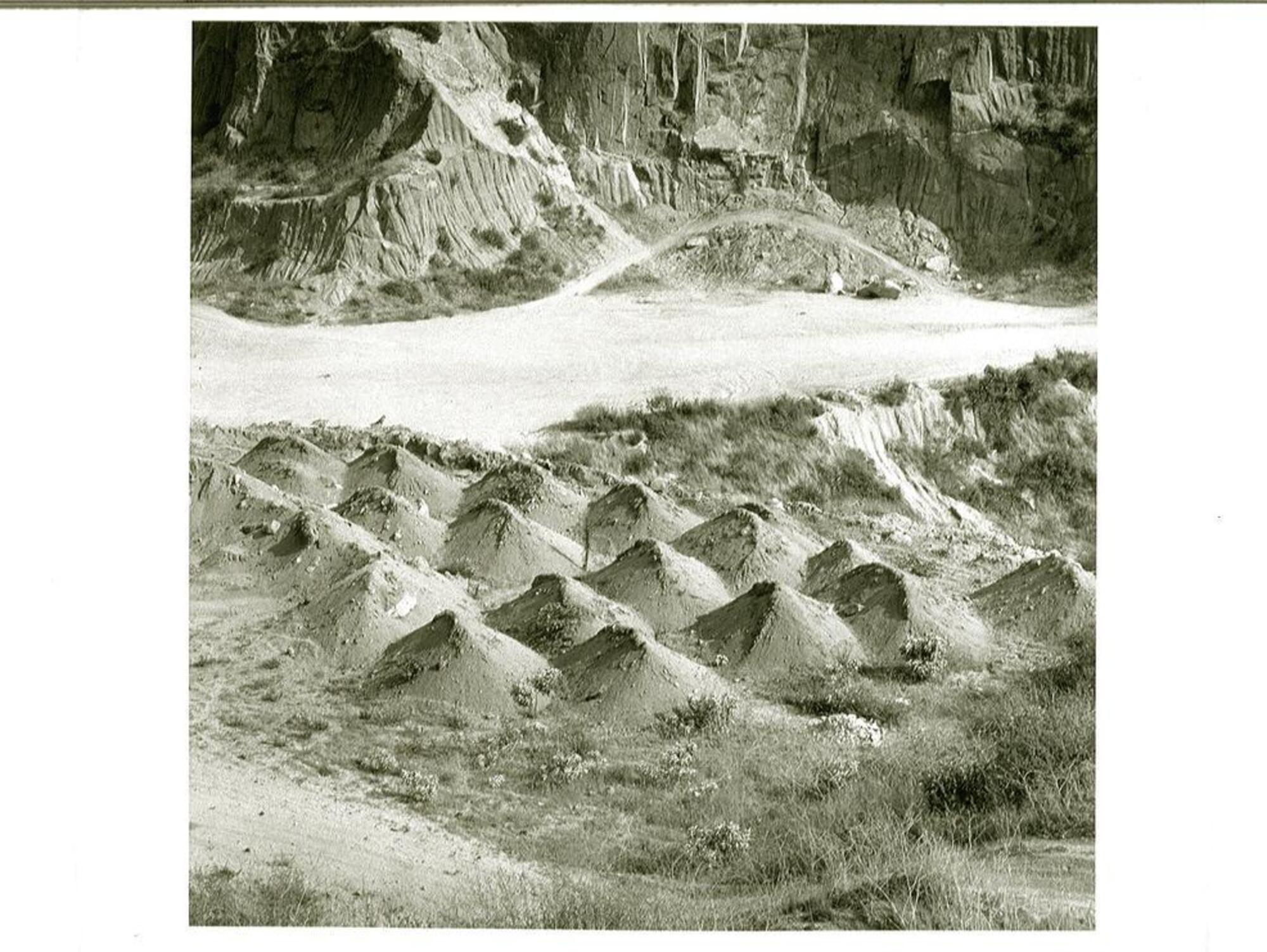 This is a black and white photograph depicting a rocky landscape scene. There are sharp cliff walls, striated with grooves, and two dirt roads with tire tracks. In the center portion, there is a grouping of seventeen piles of dirt, all of a similar size, and scrub like vegetation. The viewpoint is from above looking down.
