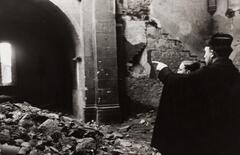 A photograph of a church destroyed by a bomb. A man in the right foreground points toward an arch.