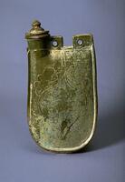 This is an earthenware flask with a tall flattened body, short neck with direct rim to one side, and two peirced, flattened lugs for a strap handle. It is incised with a floral scroll on the body and has a domed lid with tall rim and pointed finial. It is covered in a green lead glaze with iridescence and calcification. 