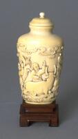 An ivory snuff bottle shaped like a vase. Carved in high relief on the surface of the snuff bottle are &nbsp;images of people, cattle, birds, and clouds. On the top of the snuff bottle is stopper. The snuff bottle is sitting on a wooden stand.