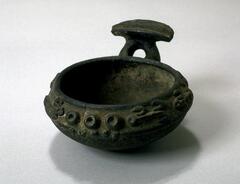 Round bowl with a raised lip and a dark patina. There is a handle on one edge with an oblong shaped projection with a large central groove. The top edge of the bowl is decorated with a pattern of raised circles and lizard motifs. 