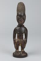 Standing male figure on a round base. The hands rest at the sides and there is a string of glass beads around the neck. The pupils of the eyes consist of metal and on each cheek is a 'V' shaped mark, while the hair is a tall oblong shape with vertical grooves. 