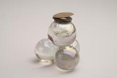 This is a clear glass inkwell with a dark metal lid. The body is composed of a large sphere balanced on three smaller spheres. It has a circular top with a flat lid.<br />