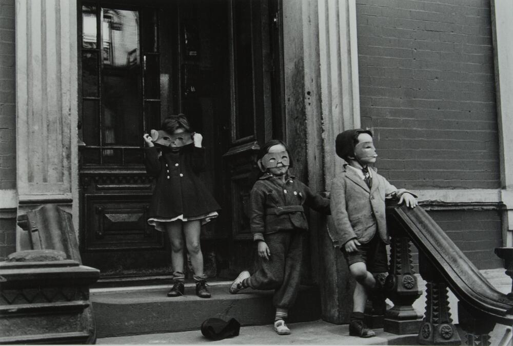 This photograph depicts three masked children standing on the steps of a browstone building in a city.