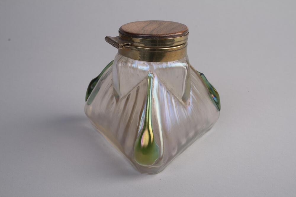 A three-sided inkwell with green iridescent colored glass. The inkwell is in a tiranglar pyramid shape, which there is a wooden cap on the top.