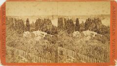 This black and white stereoscopic image features two images looking downward on a house surrounded by trees with a lake in the background and a picket fence in the lower left. This image is sepia toned mounted on a red orange background.