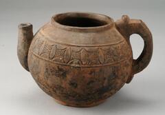 Small, round vessel in the form of a teapot. The opening has a small lip and there is a curved handle on one side of the vessel. The opposite side of the vessel has a small, curved spout. Around the top half of the vessel is a pattern of almond shapes with a scalloped edge on both sides. Between each shape is an area with small incised dots. 