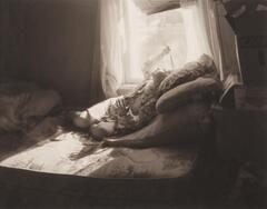 This photograph shows a young girl lying on a bed by a bright window. Her left hand is outstretched while her right holds a section of hair to the light. 