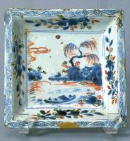 A squared porcelain dish with flat bottom, curved sides, and flat straight rim, painted in underglaze porcelain blue and overglaze red and green to create a night landscape on the base. Featuring a willow tree overlooking a body of water with a fishing boat, there is a moon in the sky, and floral sprays around the sides. Underglaze blue vine meanders around the rim. The dish is on a square footring and has gold leaf repairs.